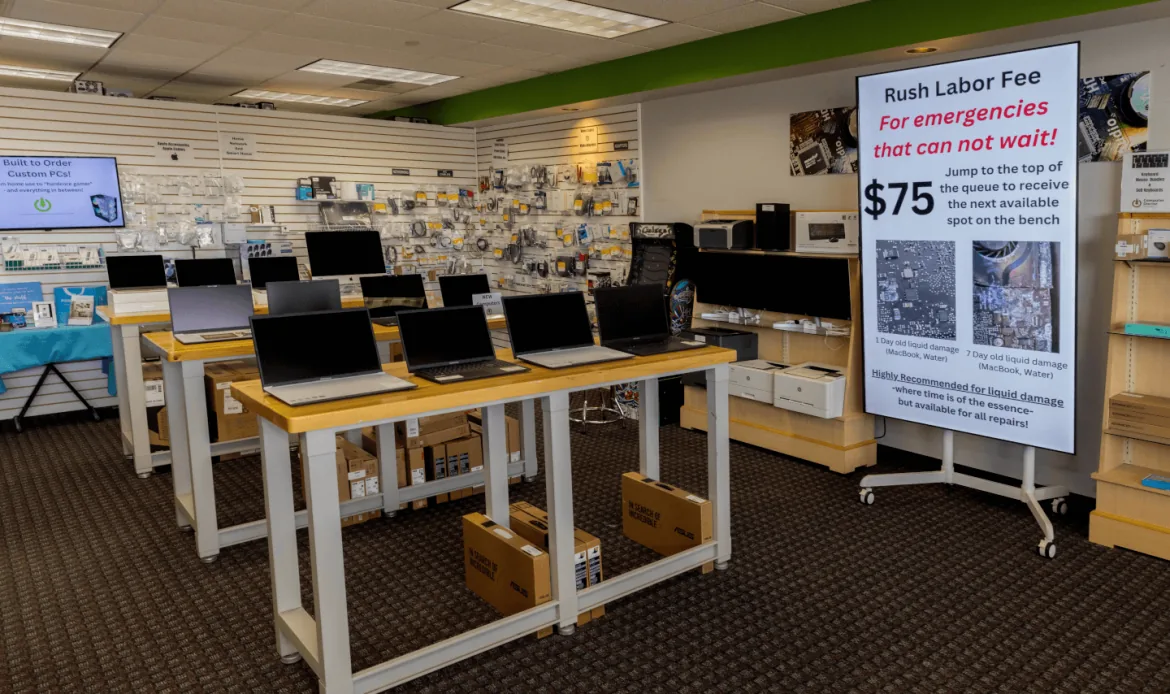 Rows of open laptops on wooden display tables on the Computer Doctor sales floor, with boxed laptops stacked underneath and a wall of accessories behind