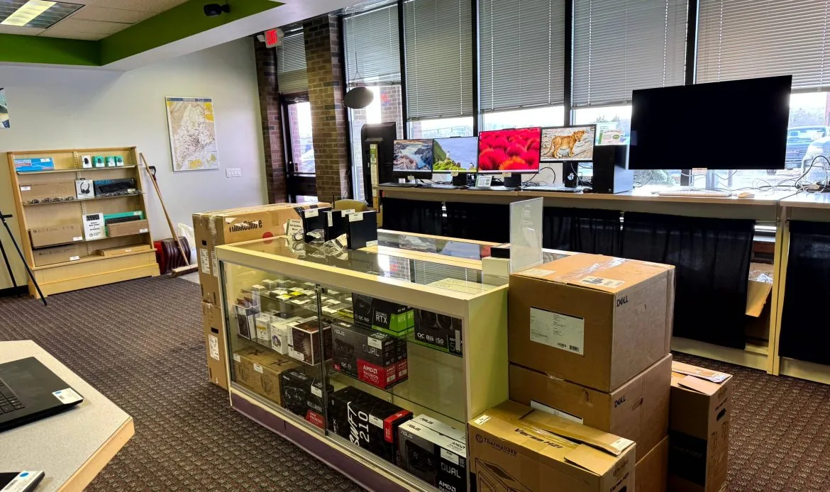 Glass display case of graphics cards and PC parts next to a stack of boxed Dell laptops, with monitors set up on the counter behind